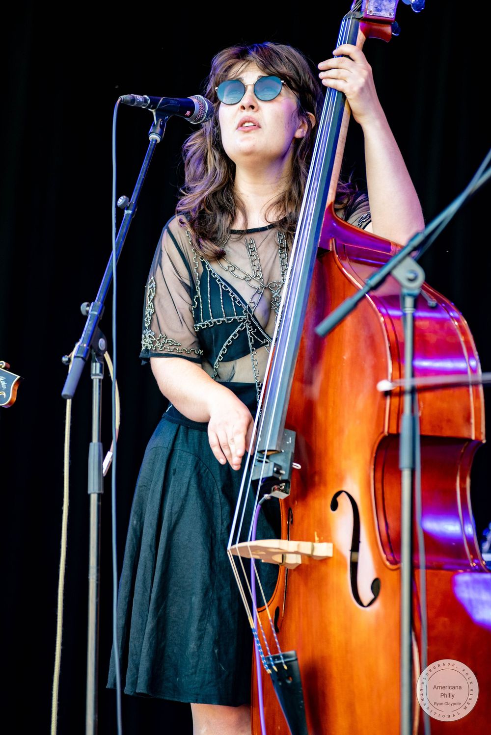 Young woman in sunglasses playing double bass on outdoor stage, wearing black skirt and sheer top.