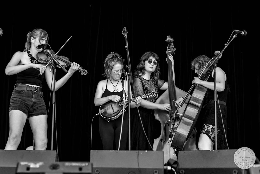 Four women performing bluegrass music on stage in black and white, playing violin, banjo, bass, and cello.