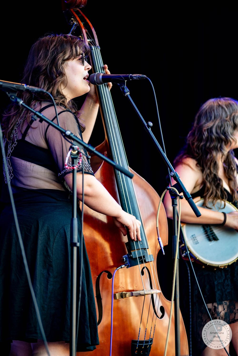 Woman playing upright bass and singing into microphone on stage at outdoor music festival.