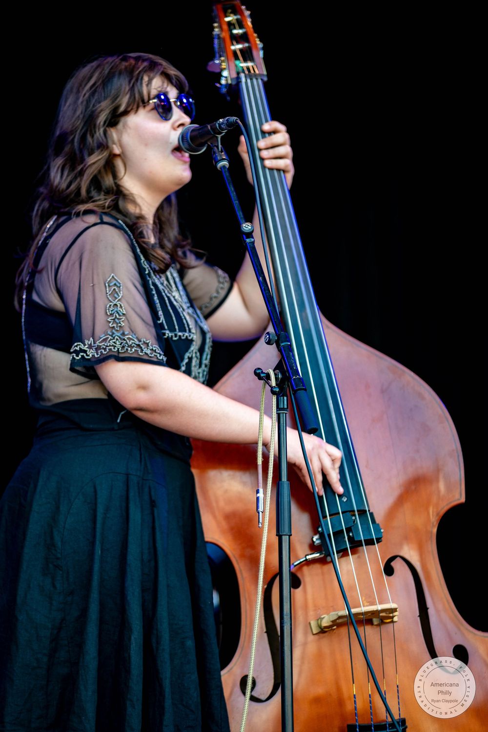 Woman playing upright bass and singing into microphone on stage, wearing sunglasses and black dress at outdoor music festival.