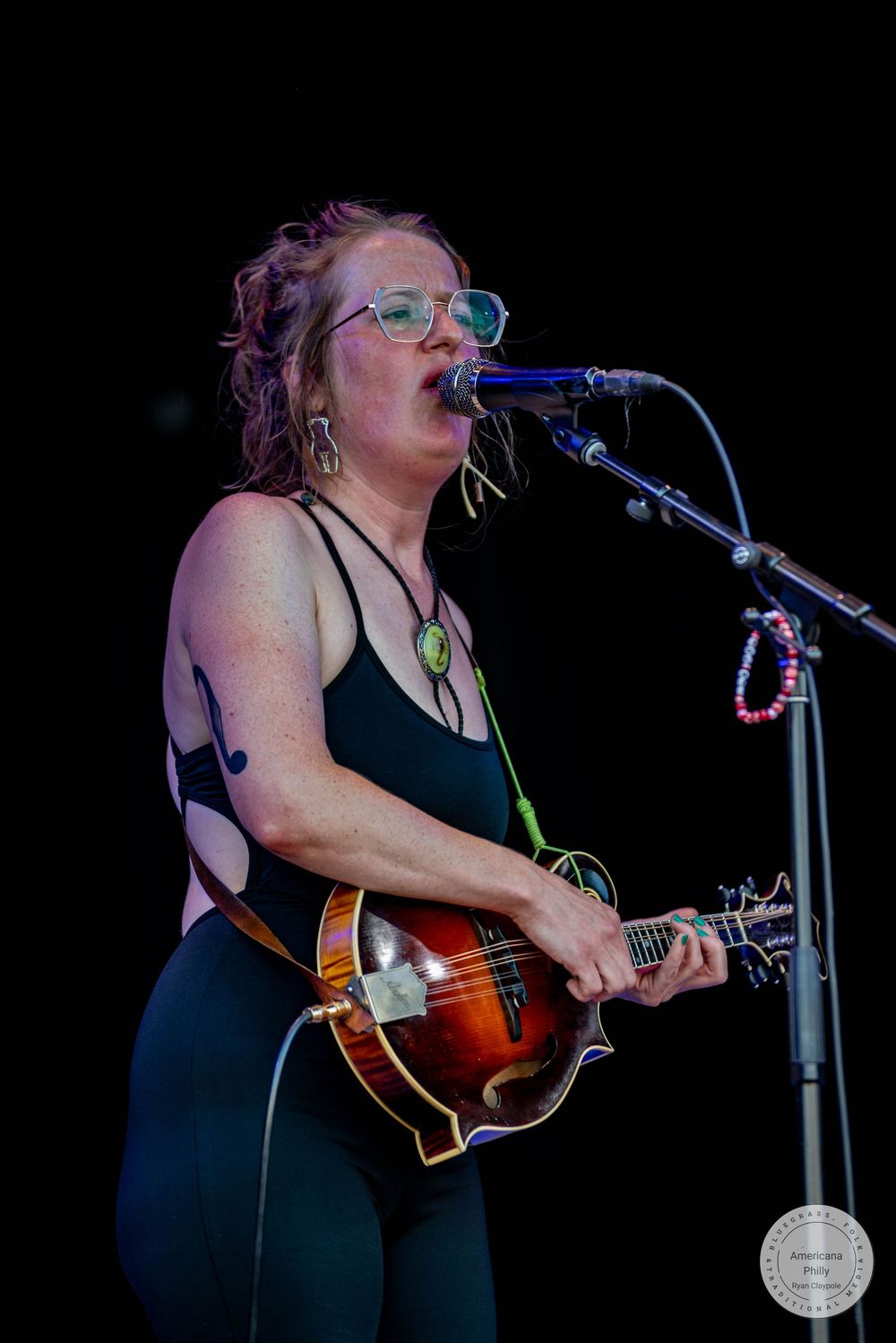 Woman playing mandolin and singing into microphone on stage, wearing black outfit and glasses at outdoor music festival.