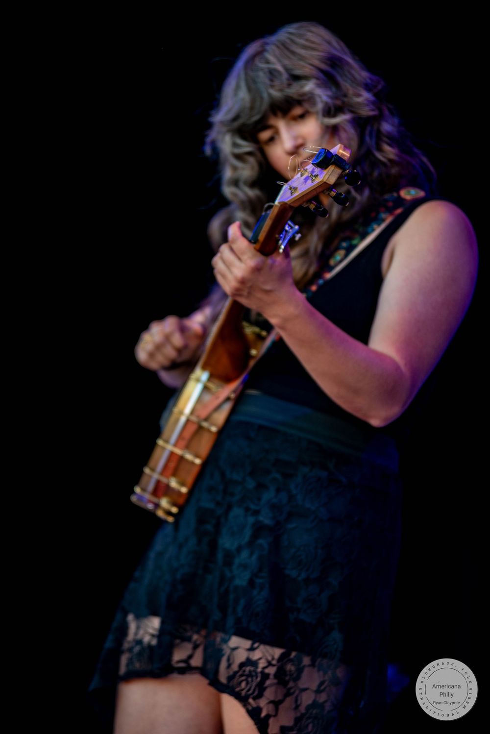 Woman in black lace dress playing ukulele on stage under purple spotlight during live performance.