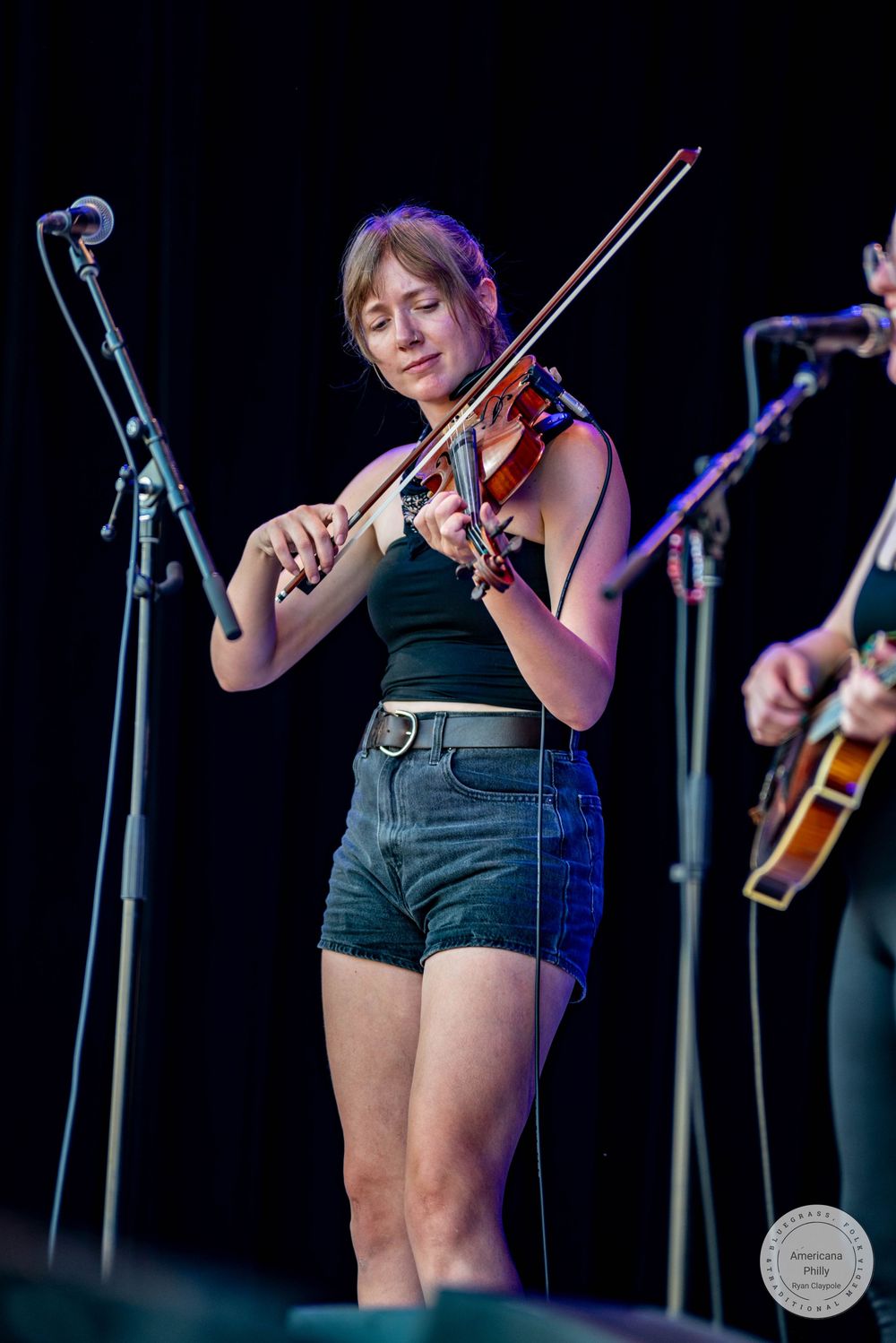 Female violinist performing on stage in black top and denim shorts at outdoor concert.