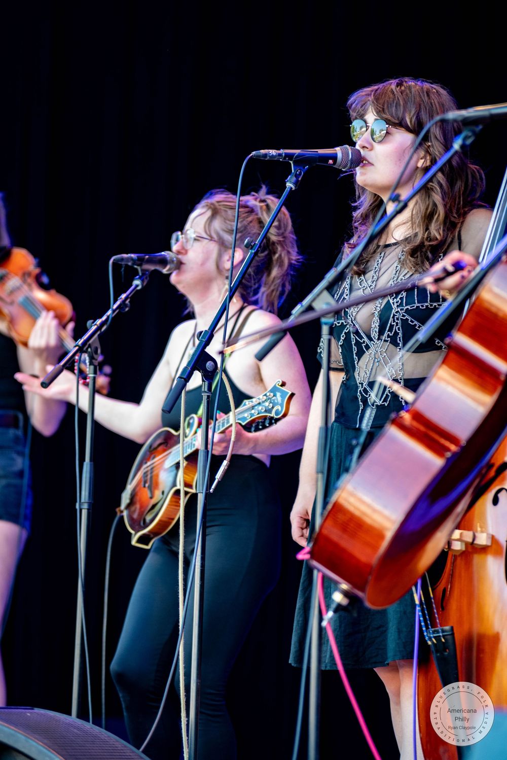 Three women perform on stage playing violin, mandolin, and cello while singing into microphones at an outdoor music festival.