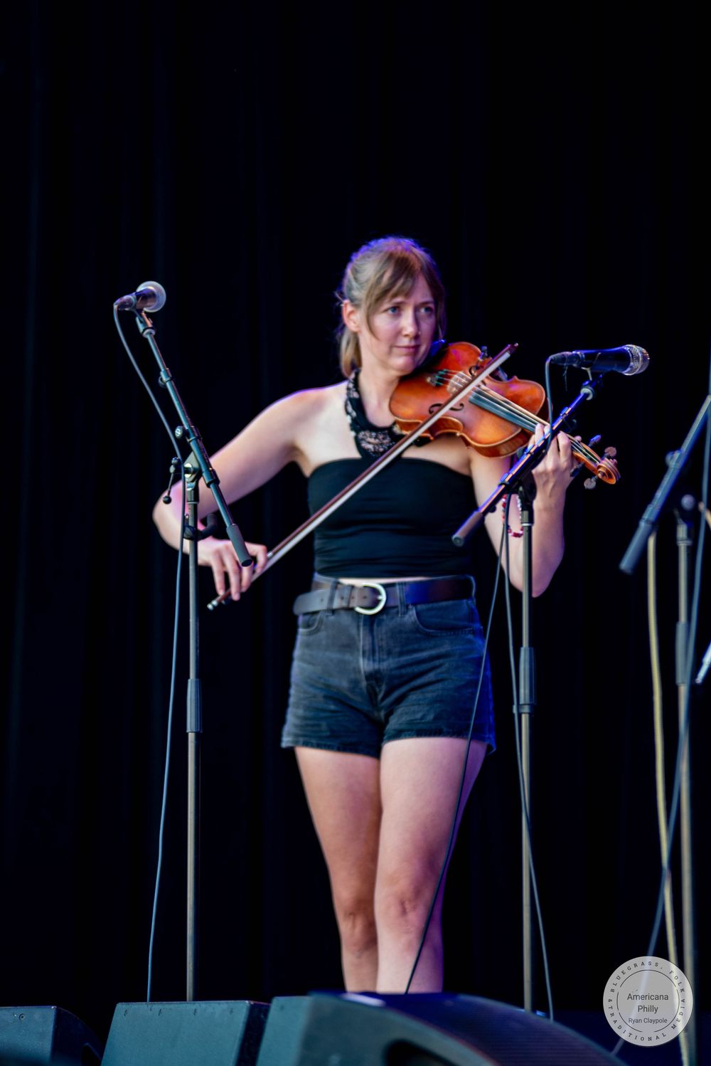 Woman playing violin on stage in black top and denim shorts at live music event.