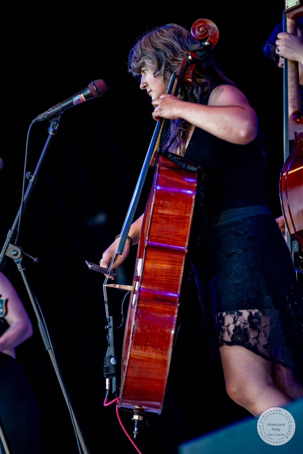 Woman playing cello on stage in black dress, performing live music concert under colorful stage lighting.