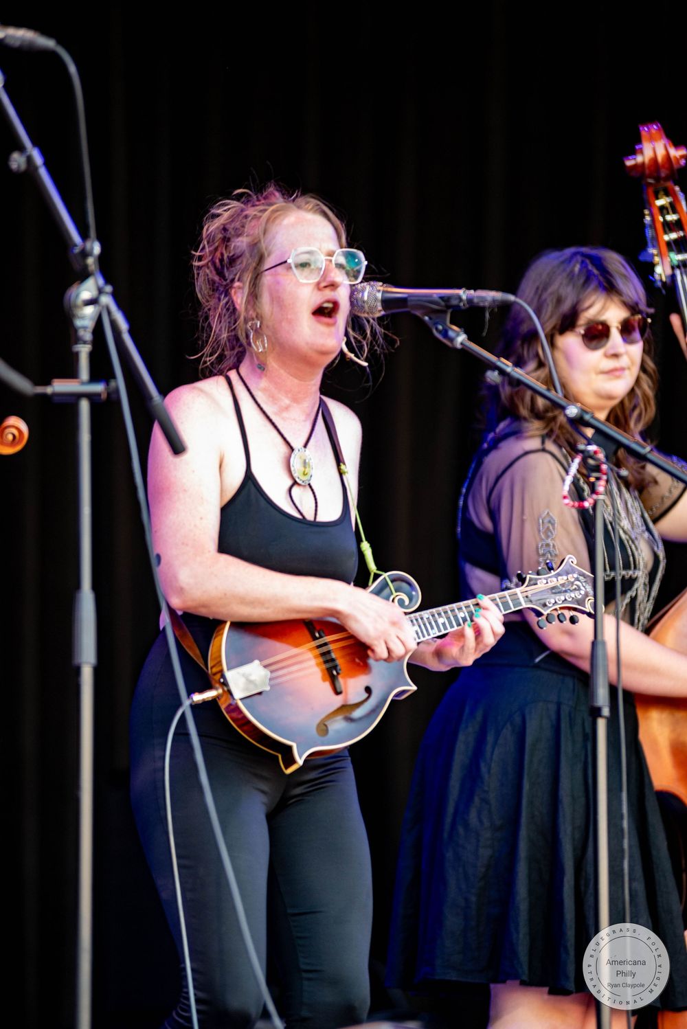 Woman playing mandolin and singing into microphone on outdoor stage during live music performance.