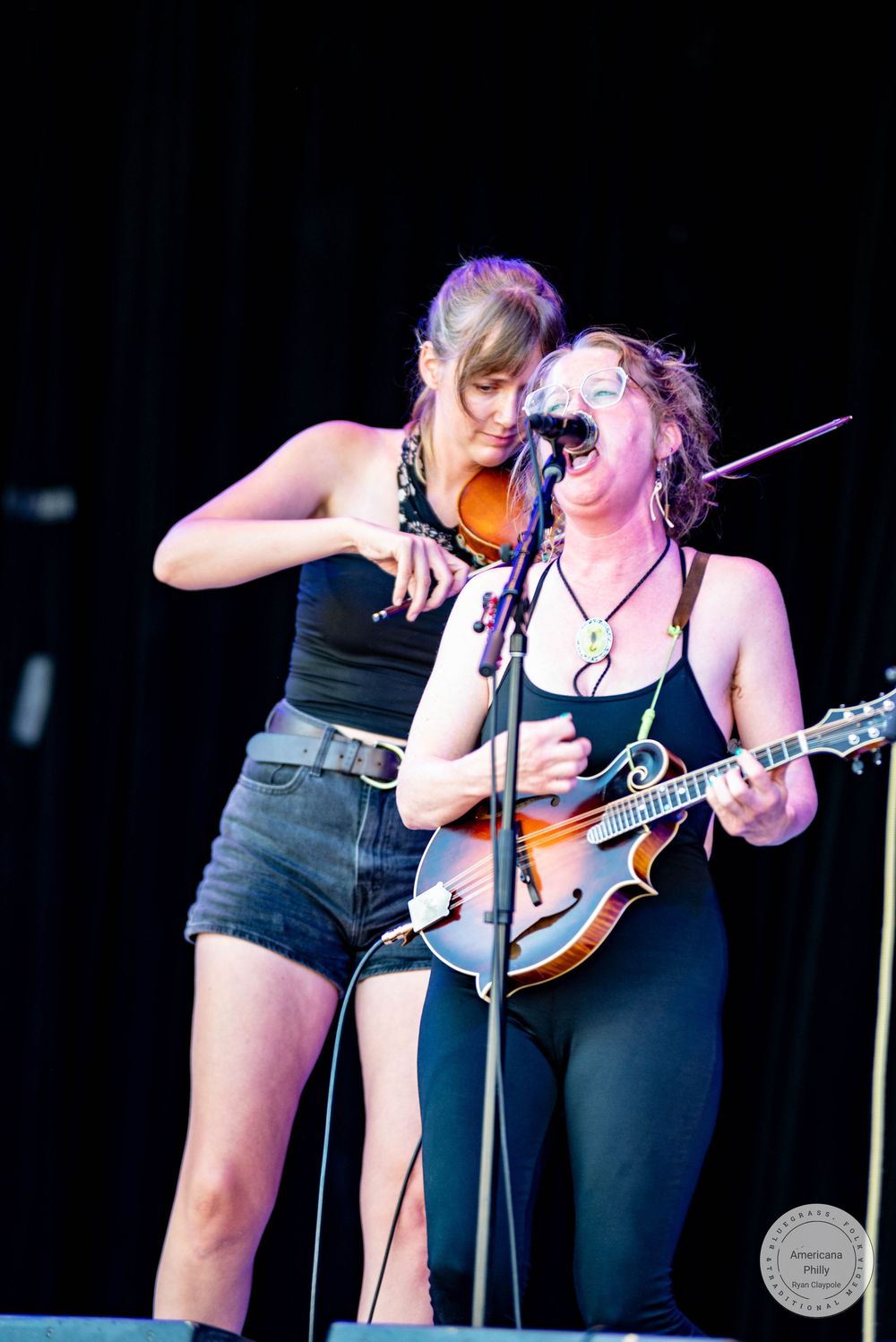Two women performing on stage, one playing mandolin at microphone, other holding violin, both wearing black outfits.