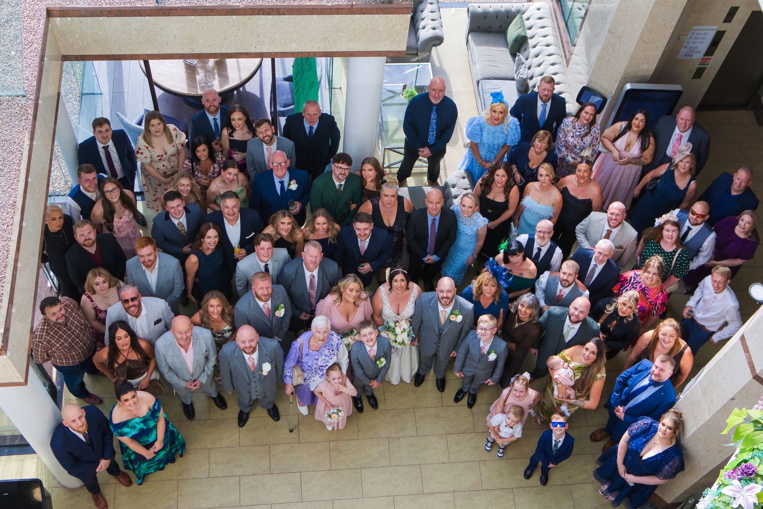 Aerial view of a large wedding group photo with bride and groom surrounded by guests in formal attire indoors.