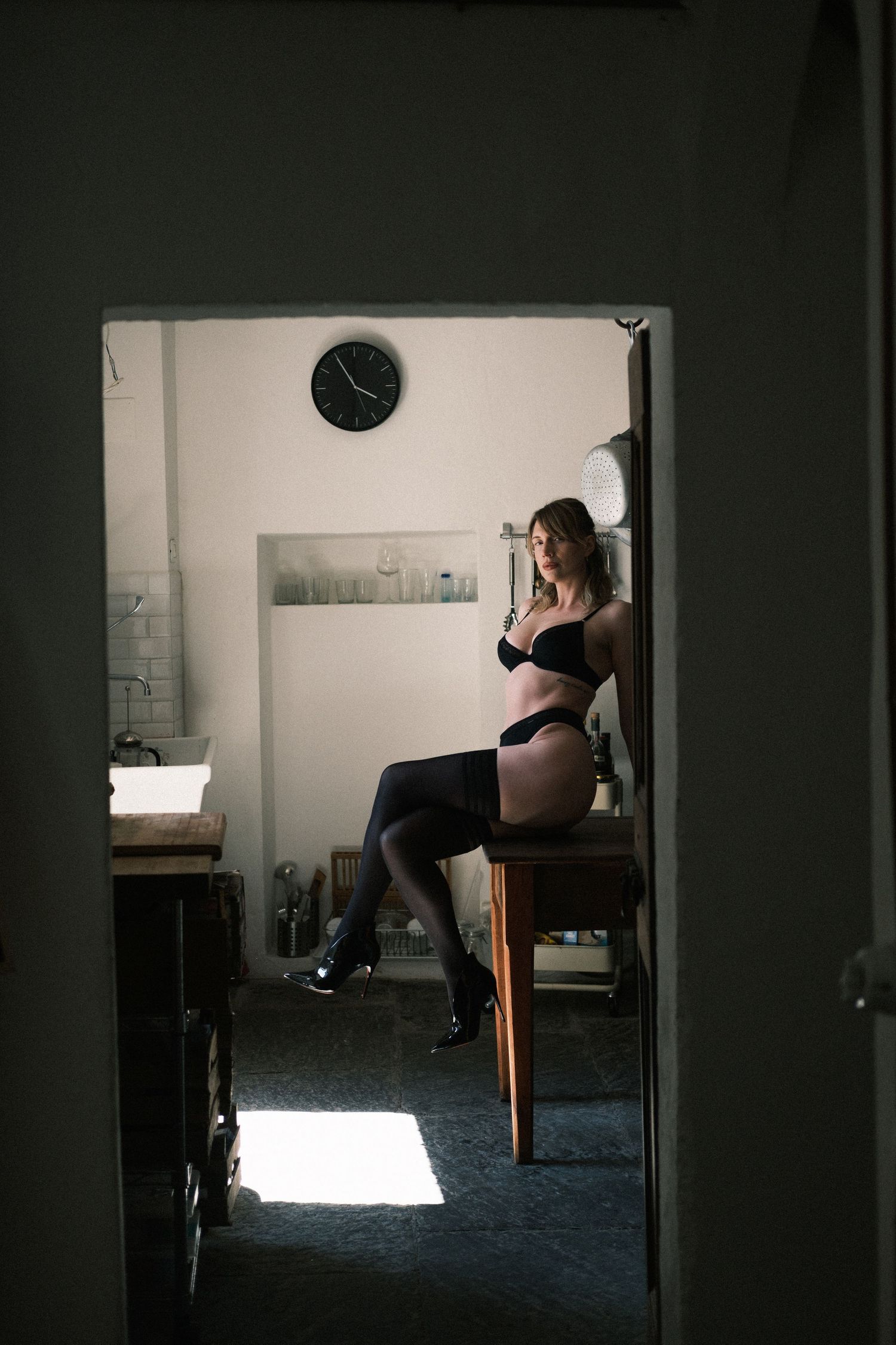 Woman in black lingerie and stockings sitting on a chair in a dimly lit room viewed through a doorway.