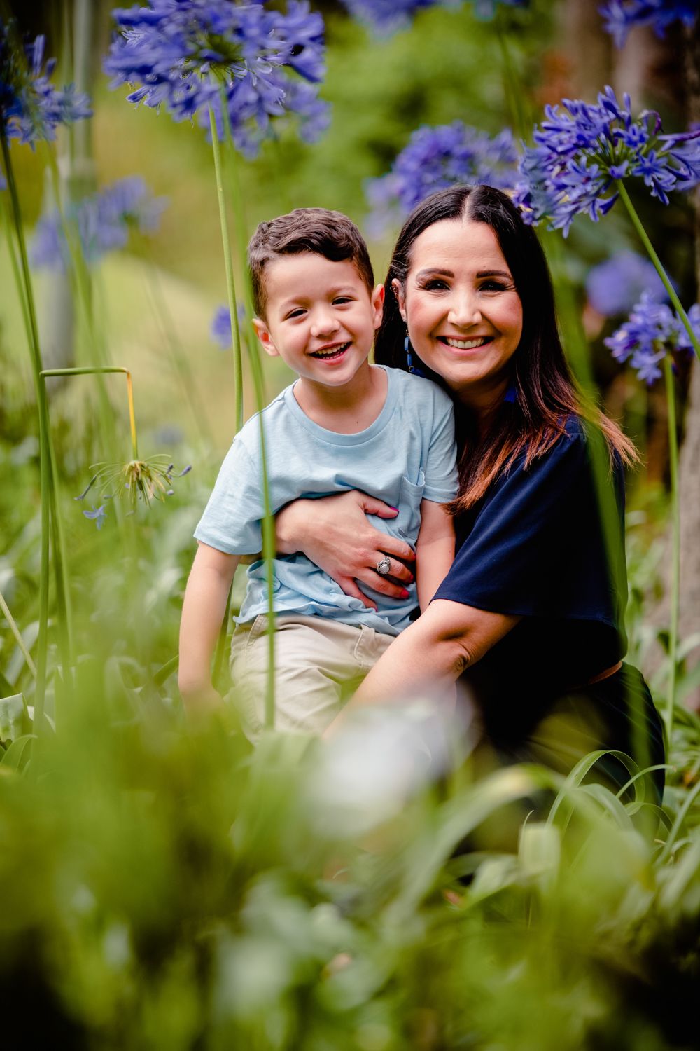 Mother hugging her son from behind amongst blue Agapanthus flowers in a garden.