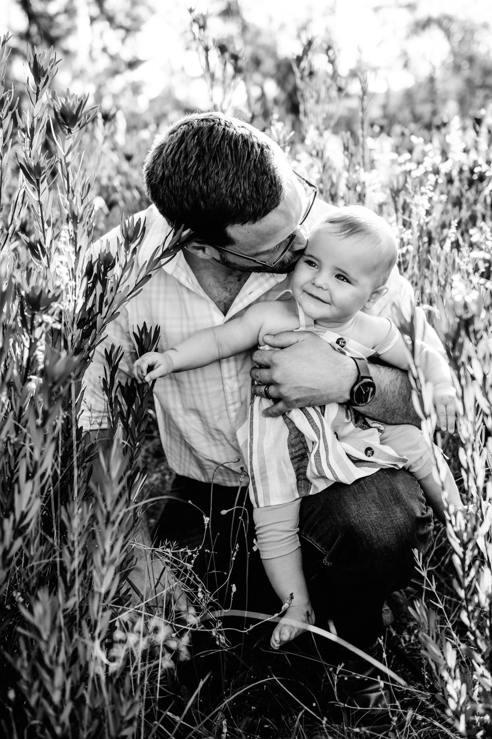 Black and white photo of a father affectionately holding his smiling toddler in a field of tall grass.