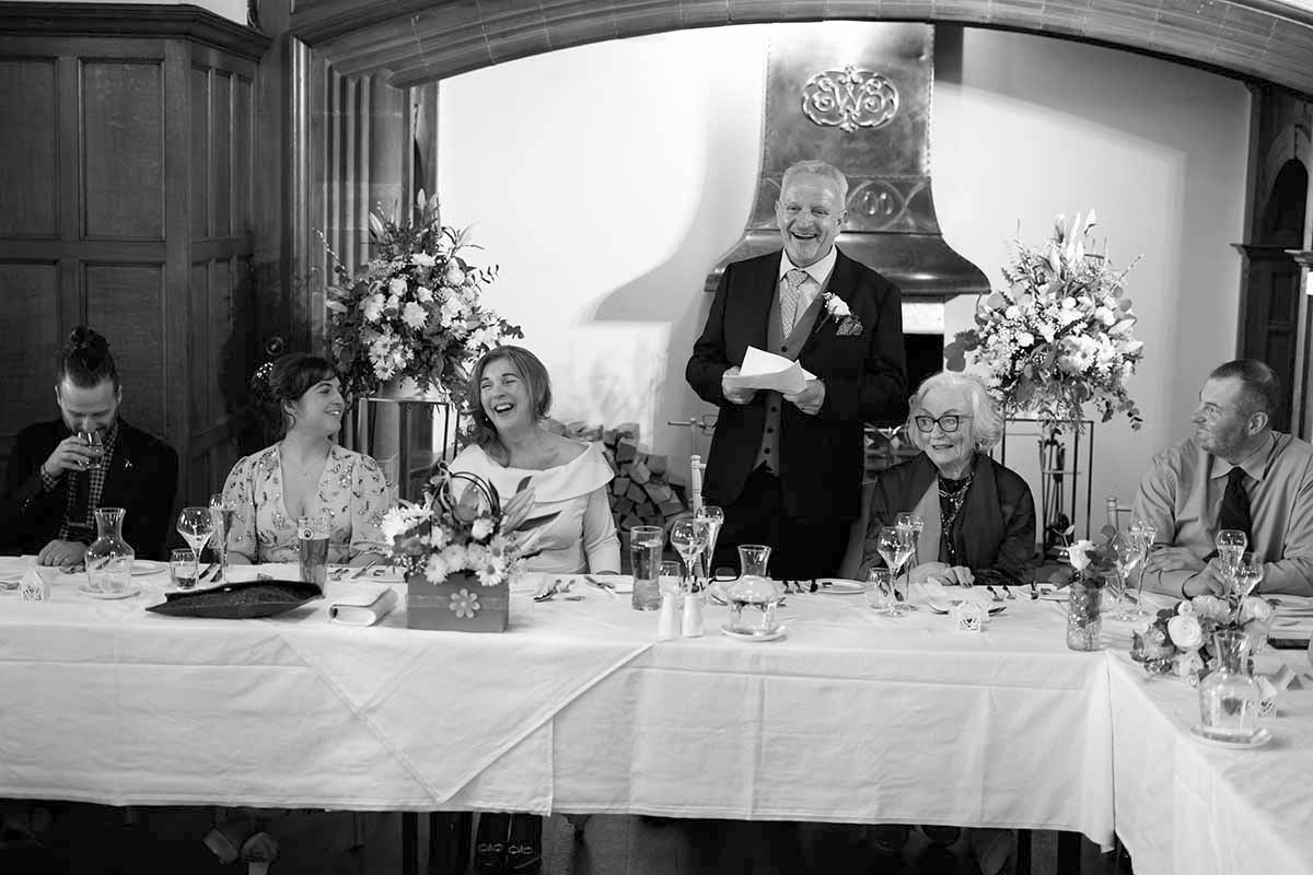 Black and white wedding reception photo showing guests at head table while man delivers speech holding paper.