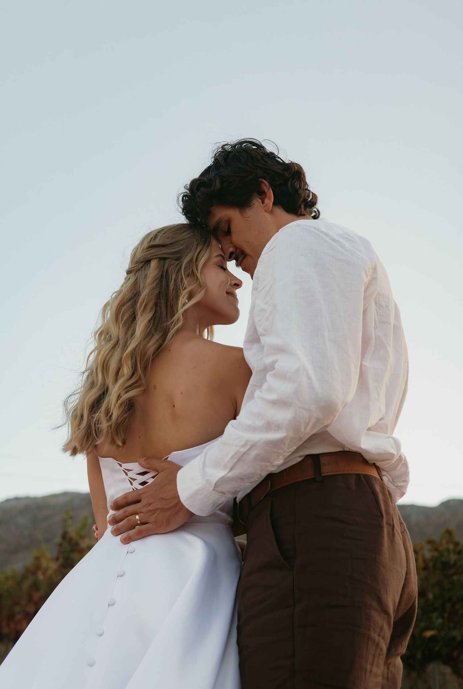 Bride in backless white dress and groom in white shirt sharing intimate moment outdoors.
