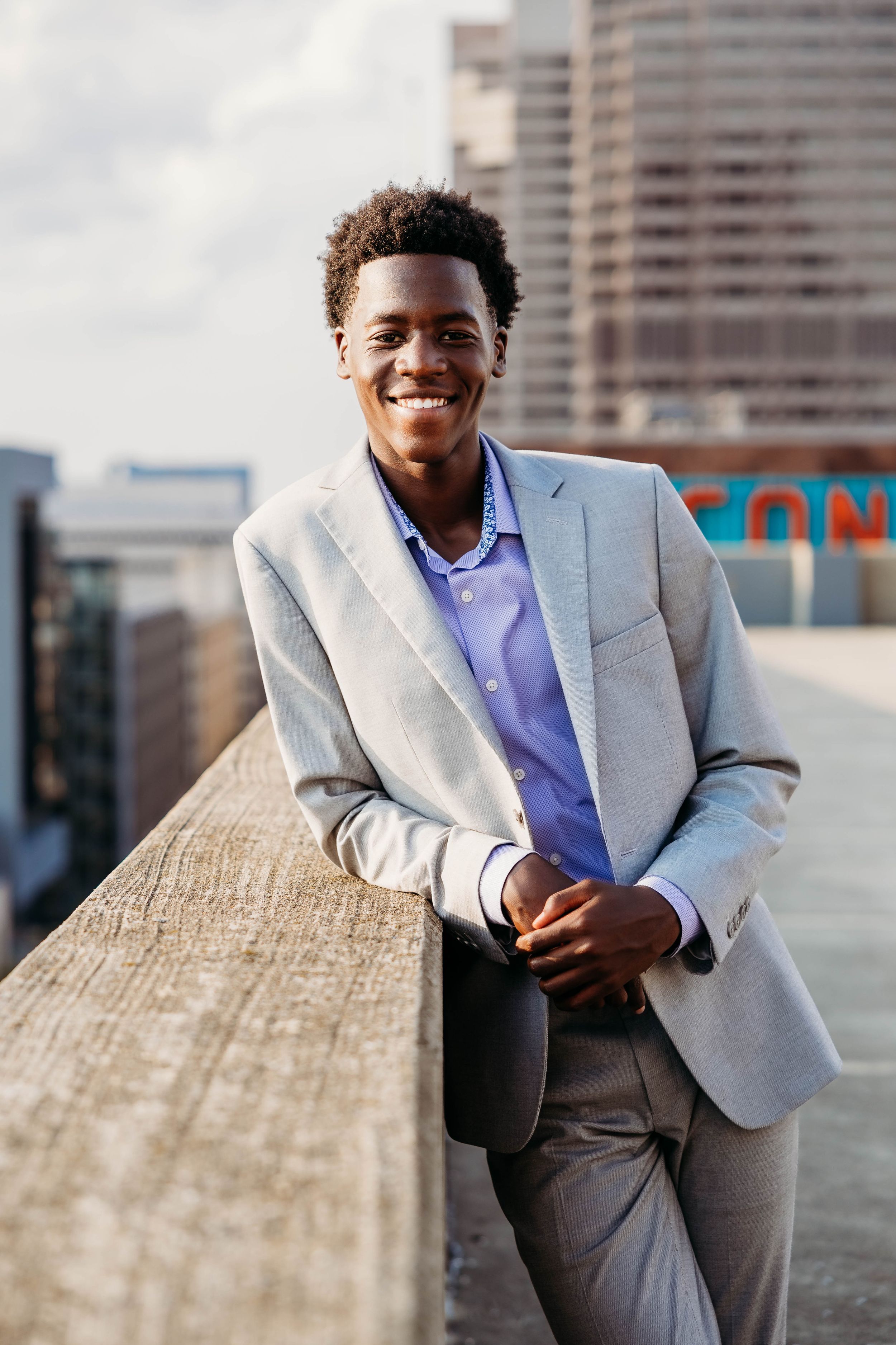 Young Black businessman in gray suit smiling on urban rooftop with city skyline background.
