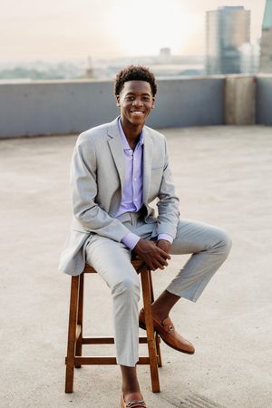 Young man in gray suit and brown loafers sits smiling on wooden stool on rooftop at sunset.
