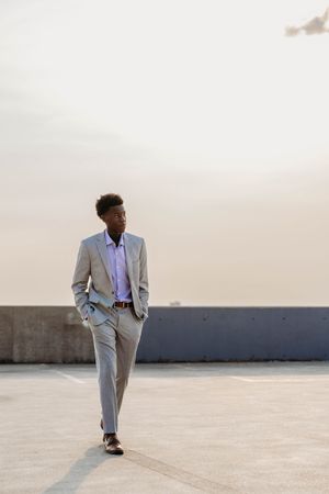 Young Black man in gray suit walking confidently on rooftop at dusk with hands in pockets.