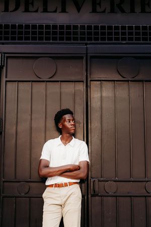 Young Black man in white polo and khaki pants stands arms crossed against dark metal delivery door in urban setting.