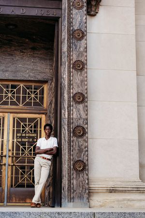 Young man in white polo and khaki pants leaning against ornate marble building entrance dated AD 1934.