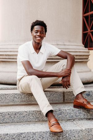 Young Black man smiling, seated on marble steps, wearing white polo, beige pants, and brown loafers.