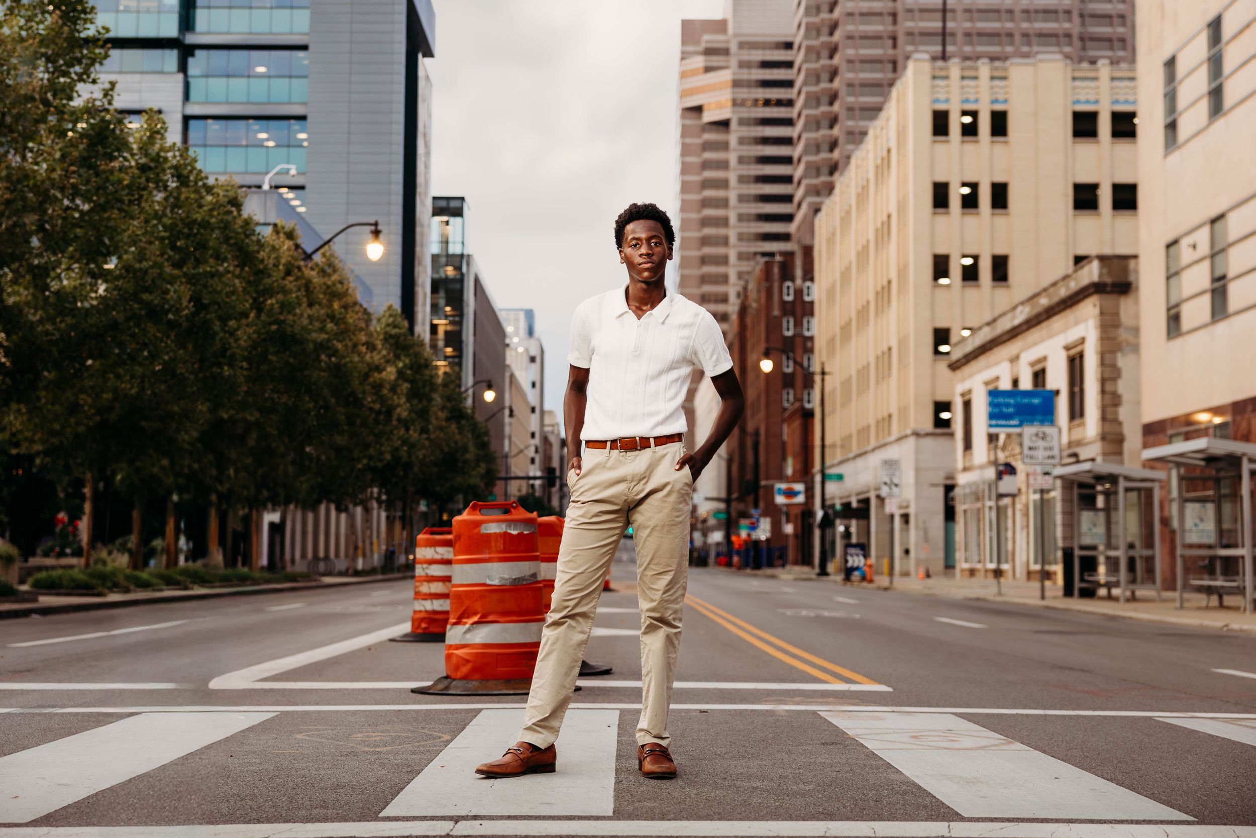 Young Black man in white polo and khakis stands confidently on a crosswalk in an urban city street.