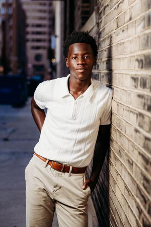 Young Black man in white zip polo and khaki pants leaning against brick wall on urban city street.