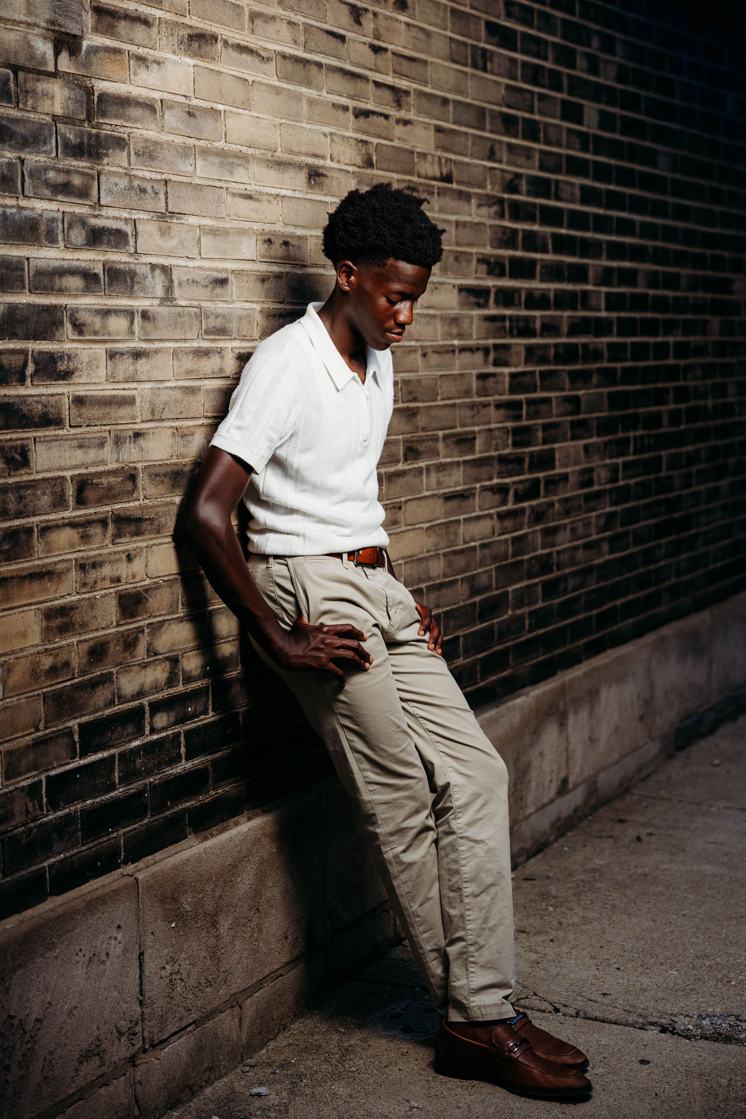 Young Black man in white polo and khaki pants leaning against a brick wall, looking down thoughtfully.