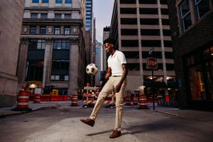 Young man skillfully juggling a soccer ball on a city street surrounded by urban buildings and construction barriers.