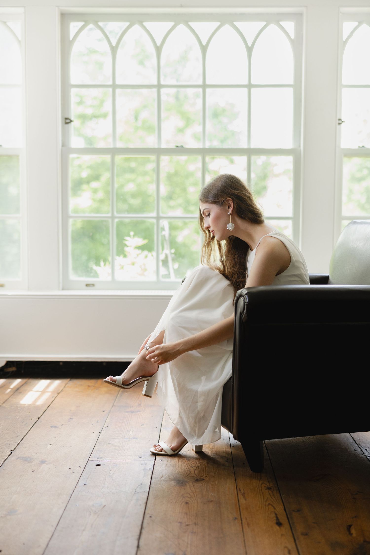 Bride in white gown adjusting heel on rustic wood floor by arched windows in sunlit room.
