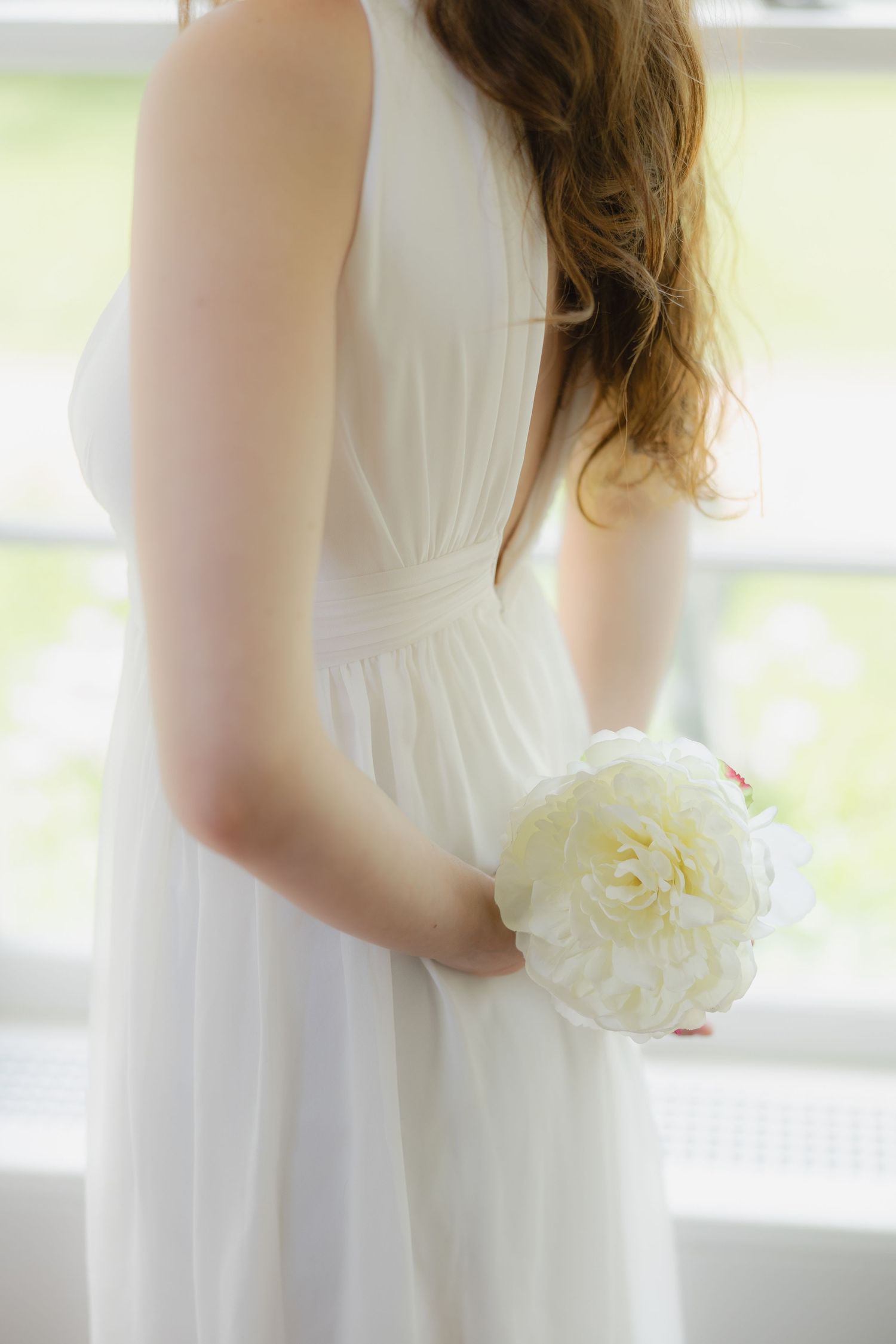 Bride in white chiffon dress holding a white peony bouquet, standing by a bright window on her wedding day.