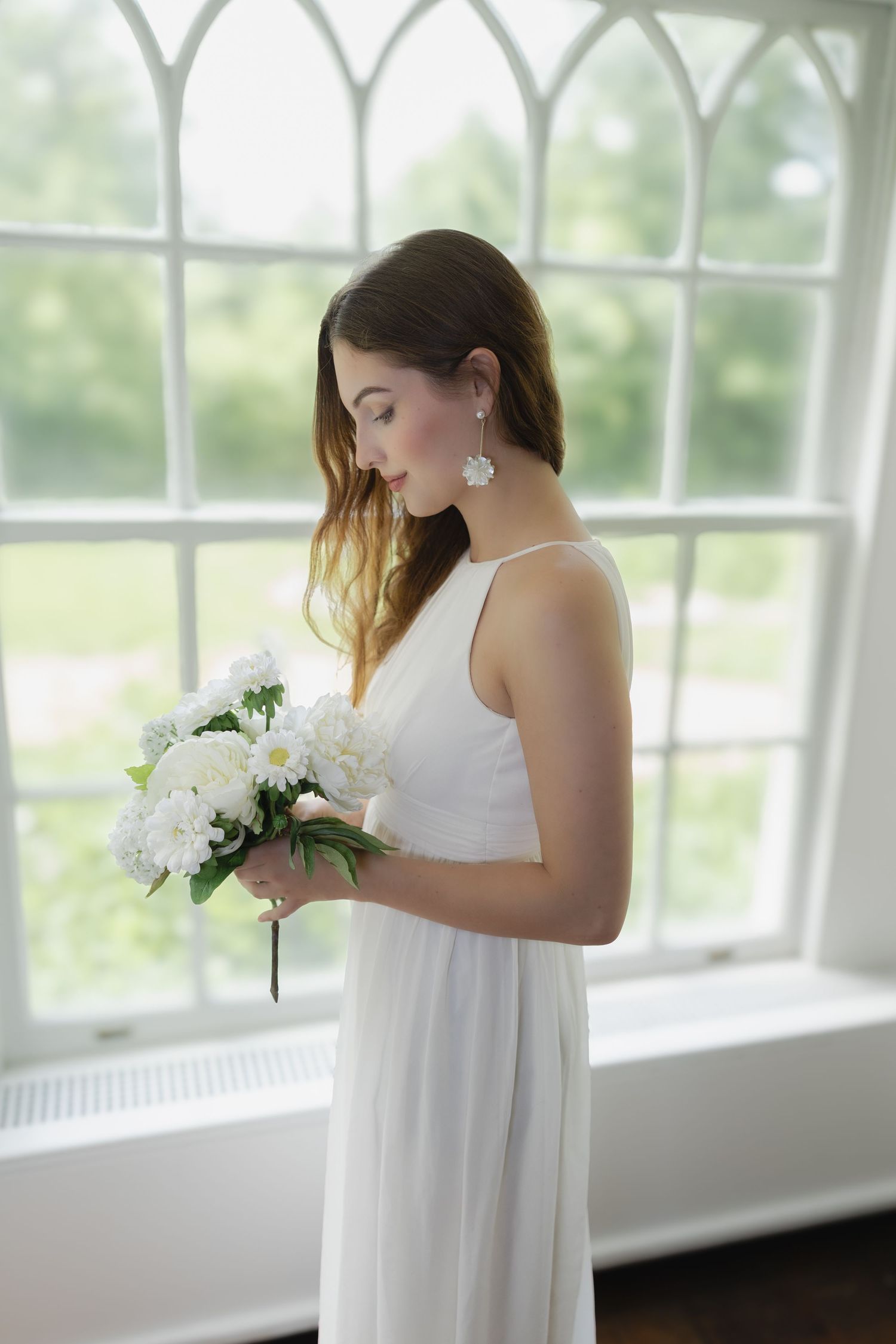 Bride in white halter gown holds white floral bouquet by arched window, soft natural light, elegant wedding portrait.