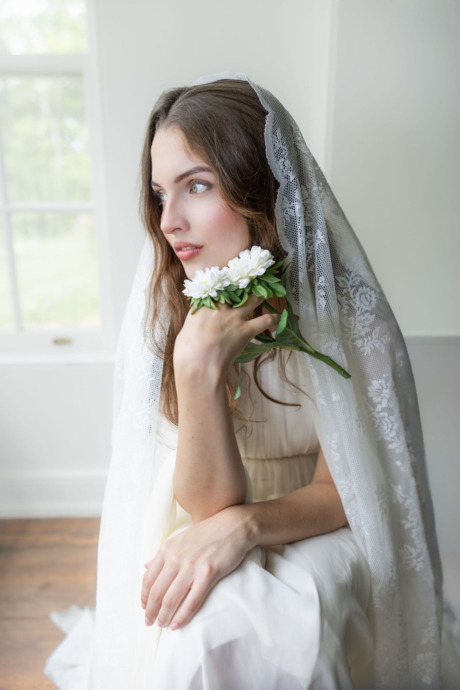 Bride in white lace veil holding white peonies, seated by window in elegant bridal gown.