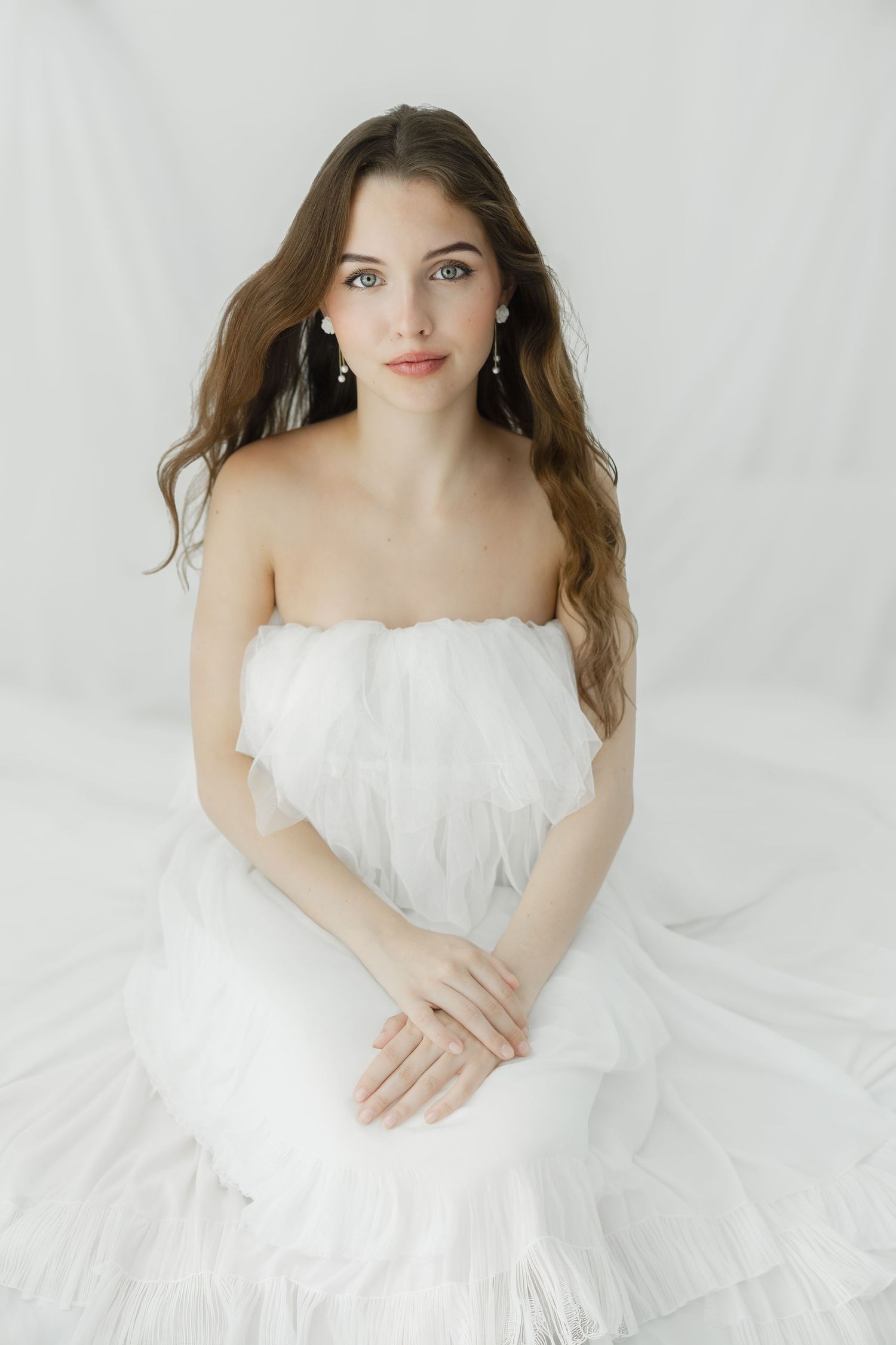 Young woman in a white strapless tulle gown with pearl earrings sits gracefully against a white background.