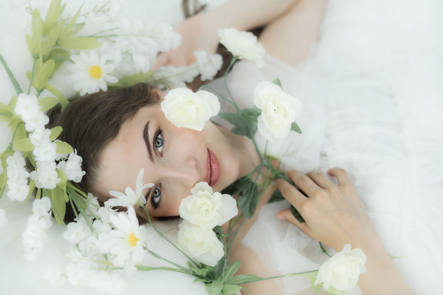 A young woman with dark hair lies among white flowers including daisies and carnations in a dreamy, ethereal setting.