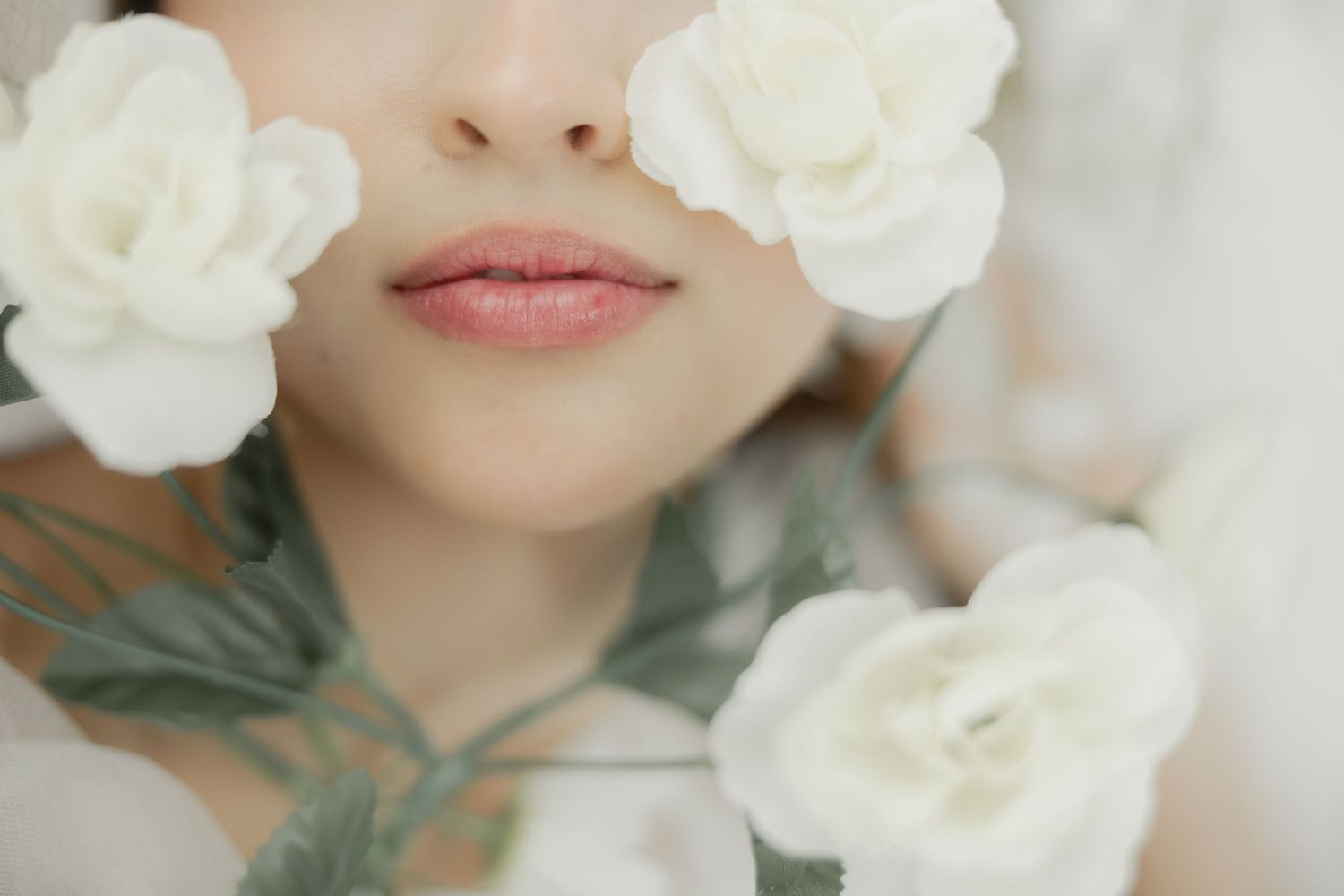 Close-up of a woman's lips surrounded by white roses and green leaves in a soft, dreamy floral portrait.