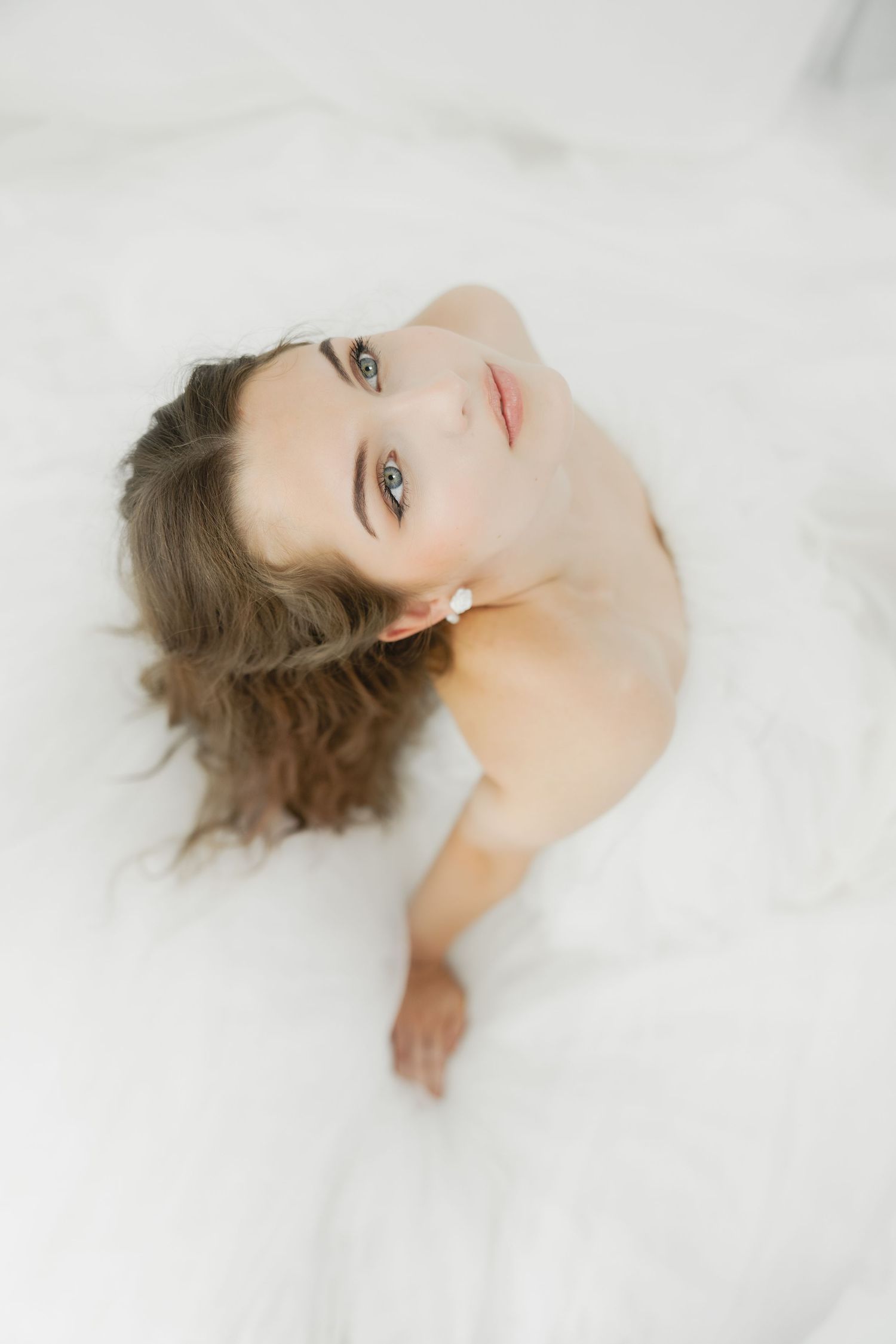 Young woman with blue eyes lying in white tulle fabric, looking up dreamily, wearing pearl earrings.