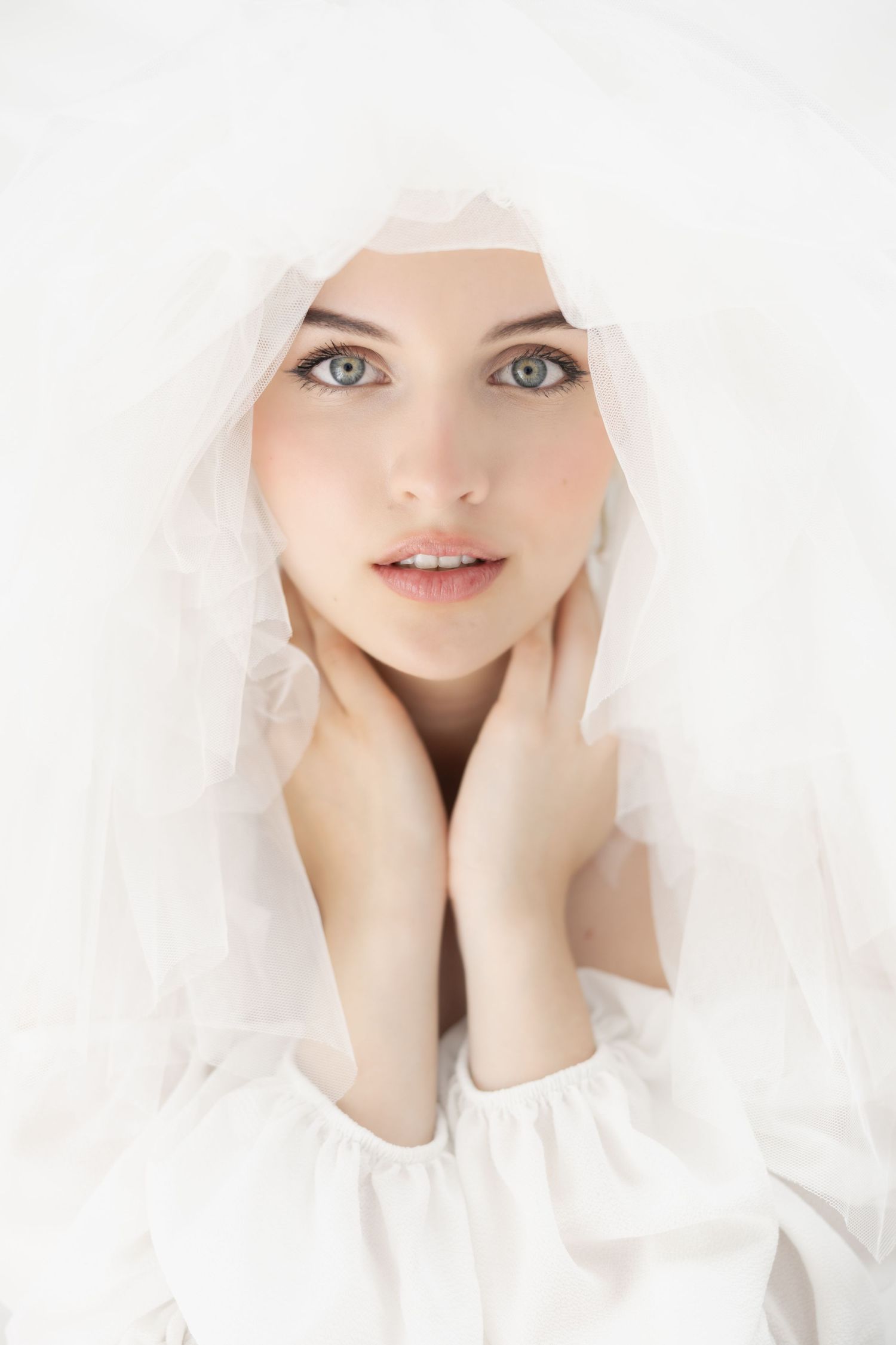Young woman with blue eyes wearing a white veil and dress, hands framing her face in an elegant bridal portrait.