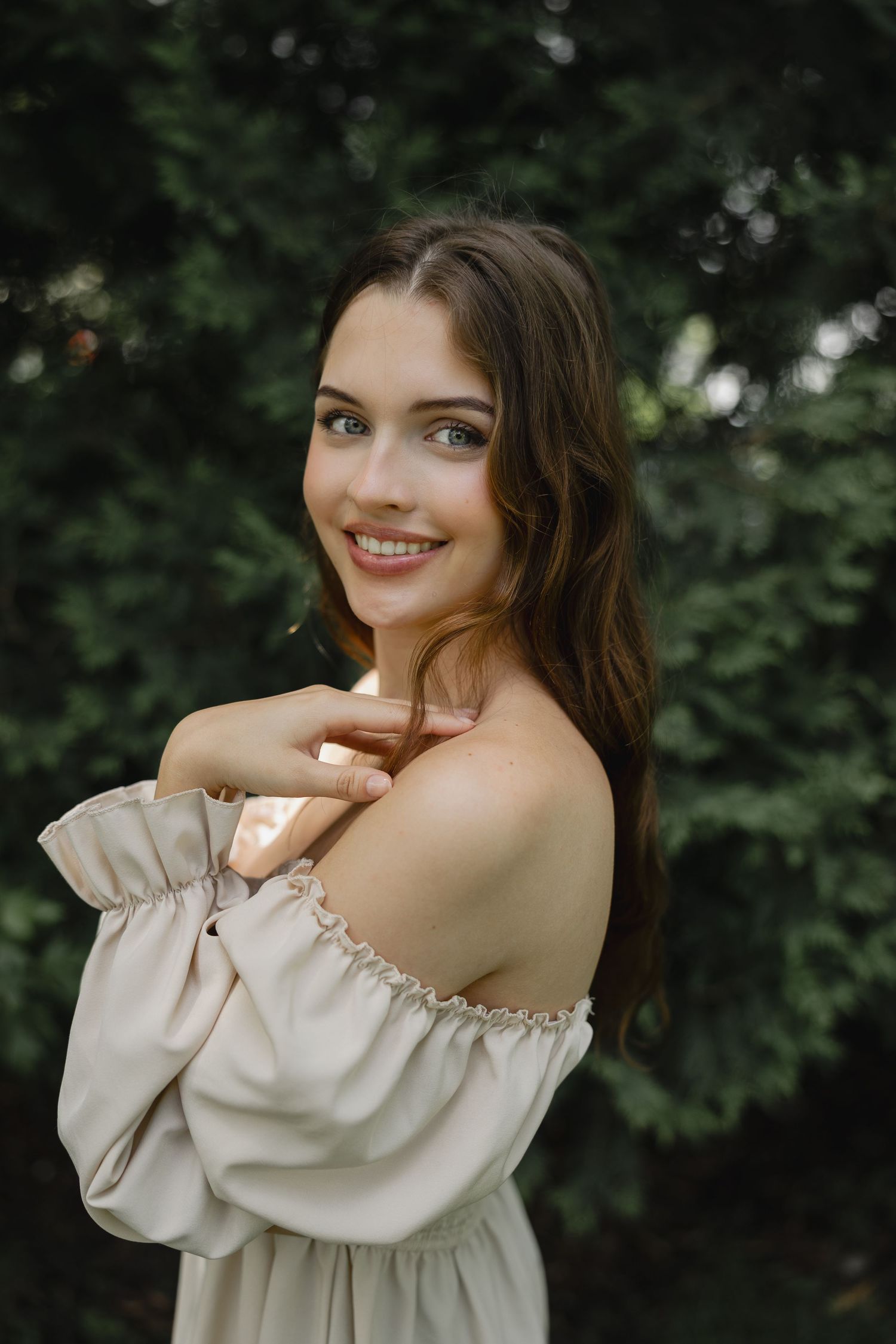 Young woman in off-shoulder beige dress smiling outdoors with lush green trees in background.