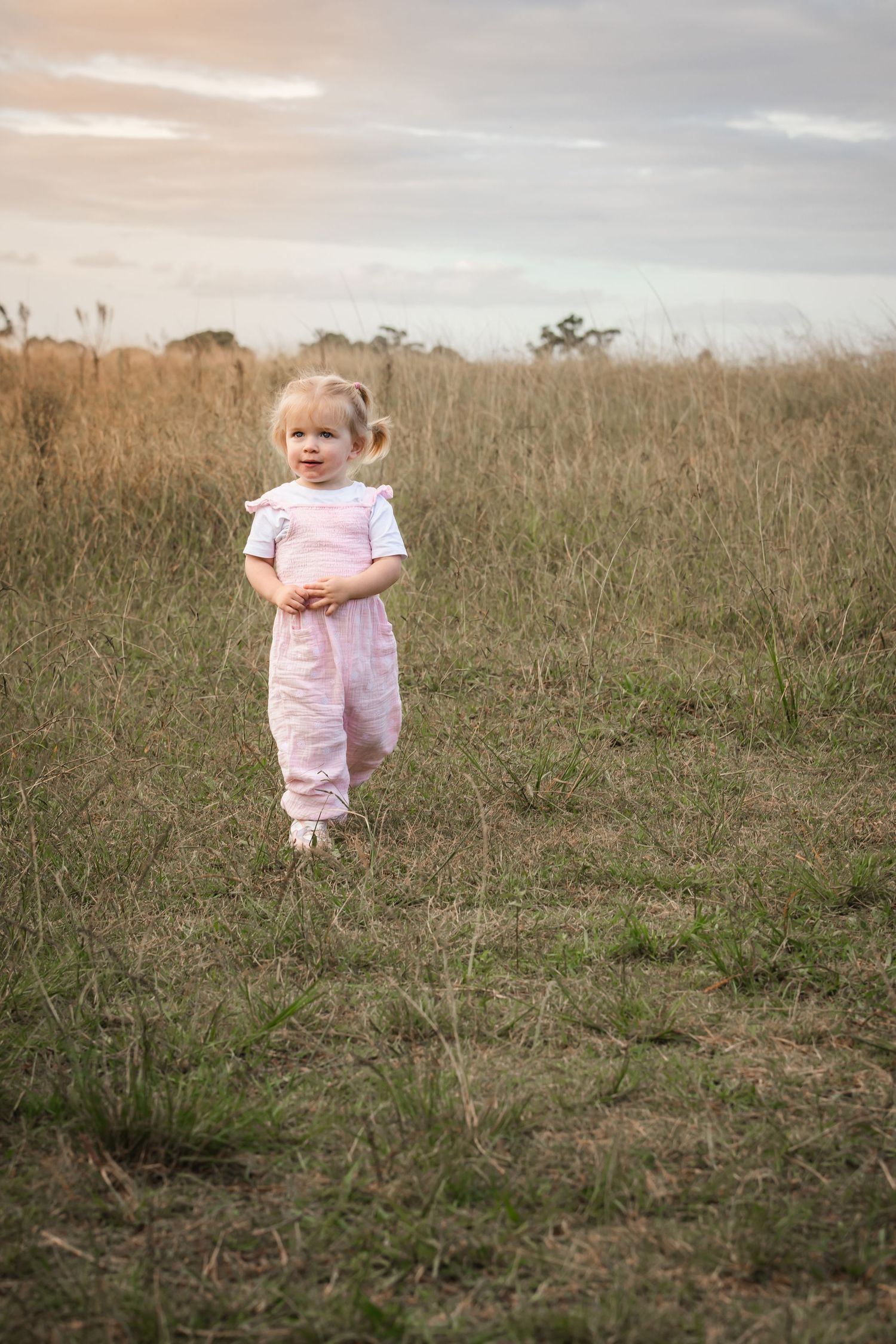 Toddler girl in pink overalls standing in a grassy field at Rouse Hill Regional Park at dusk.