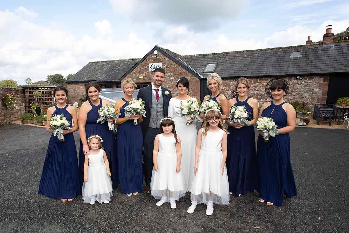Wedding party poses outdoors, bride and groom flanked by bridesmaids in navy gowns and flower girls in white dresses.