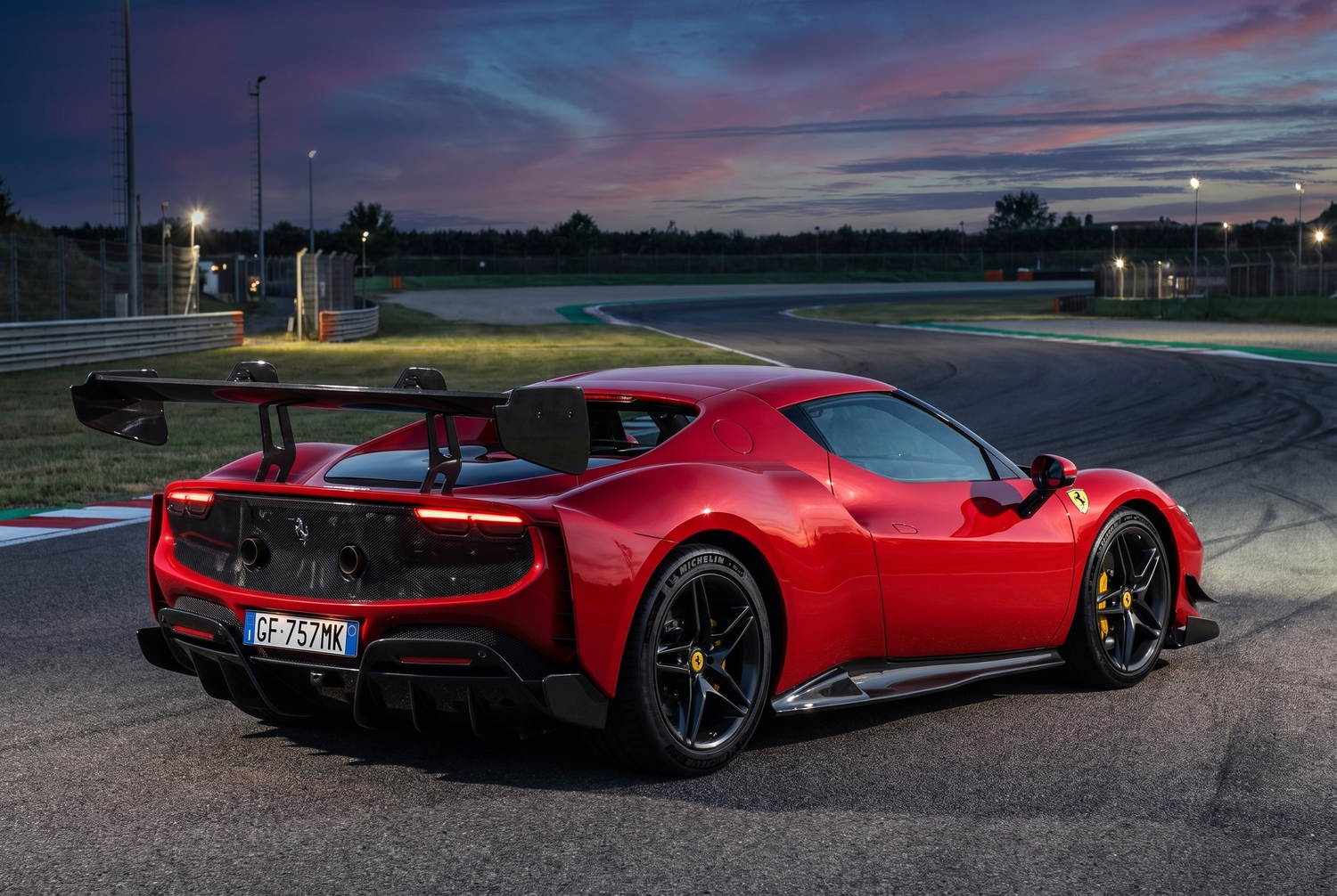 Red Ferrari supercar rear view on a race track at dusk, featuring large spoiler and aggressive aerodynamic bodywork.