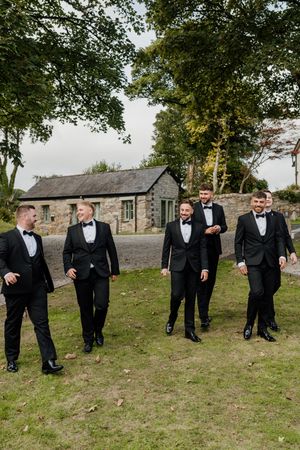 Six groomsmen in black tuxedos walking and laughing together outdoors near a stone building on a wedding day.