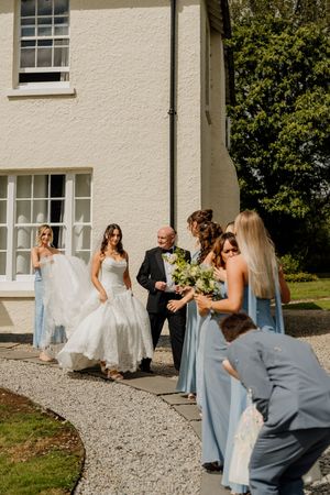 Bride in white gown walks with bridesmaids in blue dresses and groom in tuxedo outside a white country house.