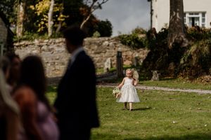 Young flower girl in white dress walks across lawn during outdoor wedding ceremony.