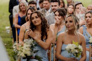 Emotional bridesmaids in light blue dresses holding white bouquets during an outdoor wedding ceremony.