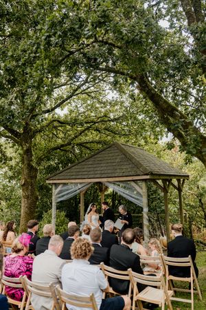 Outdoor wedding ceremony under a wooden gazebo surrounded by lush green trees with guests seated on wooden chairs.
