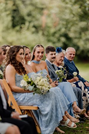 Wedding guests in blue dresses and suits seated outdoors, smiling during a garden ceremony with floral bouquets.