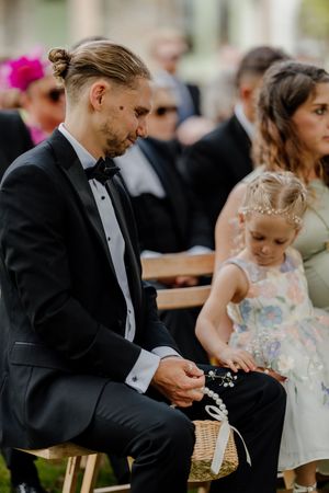 Man in tuxedo sits with young flower girl at outdoor wedding ceremony.