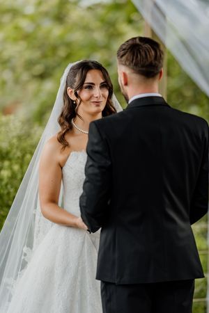 Bride in lace gown and veil holds hands with groom in black suit during outdoor wedding ceremony.