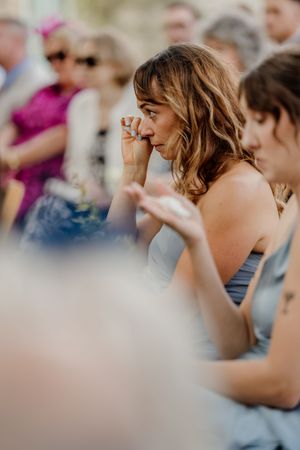 Emotional wedding guest wiping tears during outdoor ceremony, wearing blue dress.